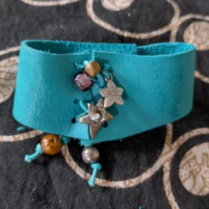 Turquoise leather cuff bracelet with silver star and flower charms and colorful beads on a patterned dark background.