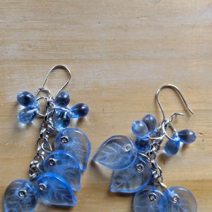 Blue glass leaf-shaped earrings with dangling round beads on metal chains, laid on a light wood surface